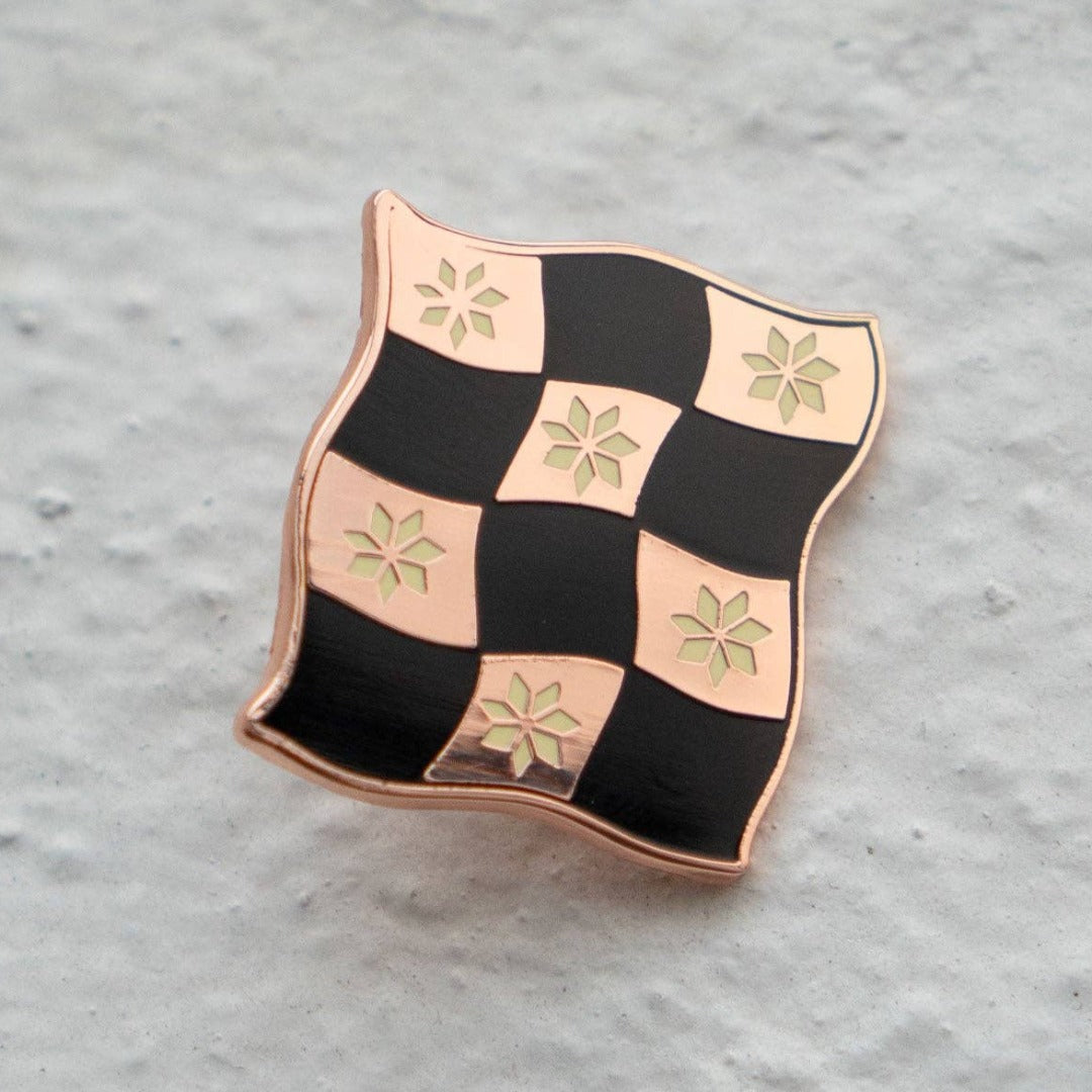 Rose gold-toned pin shaped like a checkered flag with black and white squares. Each square has a small, white flower design in the center.