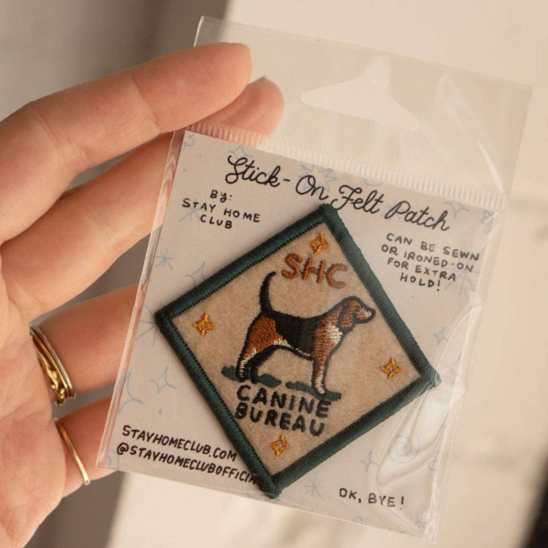 A diamond-shaped embroidered patch with a green border. It features a brown and black beagle standing facing left, with the text "SHC" above and "CANINE BUREAU" below the dog. The patch has four small stars at the corners.