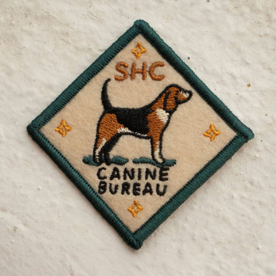A diamond-shaped embroidered patch with a green border. It features a brown and black beagle standing facing left, with the text "SHC" above and "CANINE BUREAU" below the dog. The patch has four small stars at the corners.