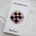 Rose gold-toned pin shaped like a checkered flag with black and white squares. Each square has a small, white flower design in the center.