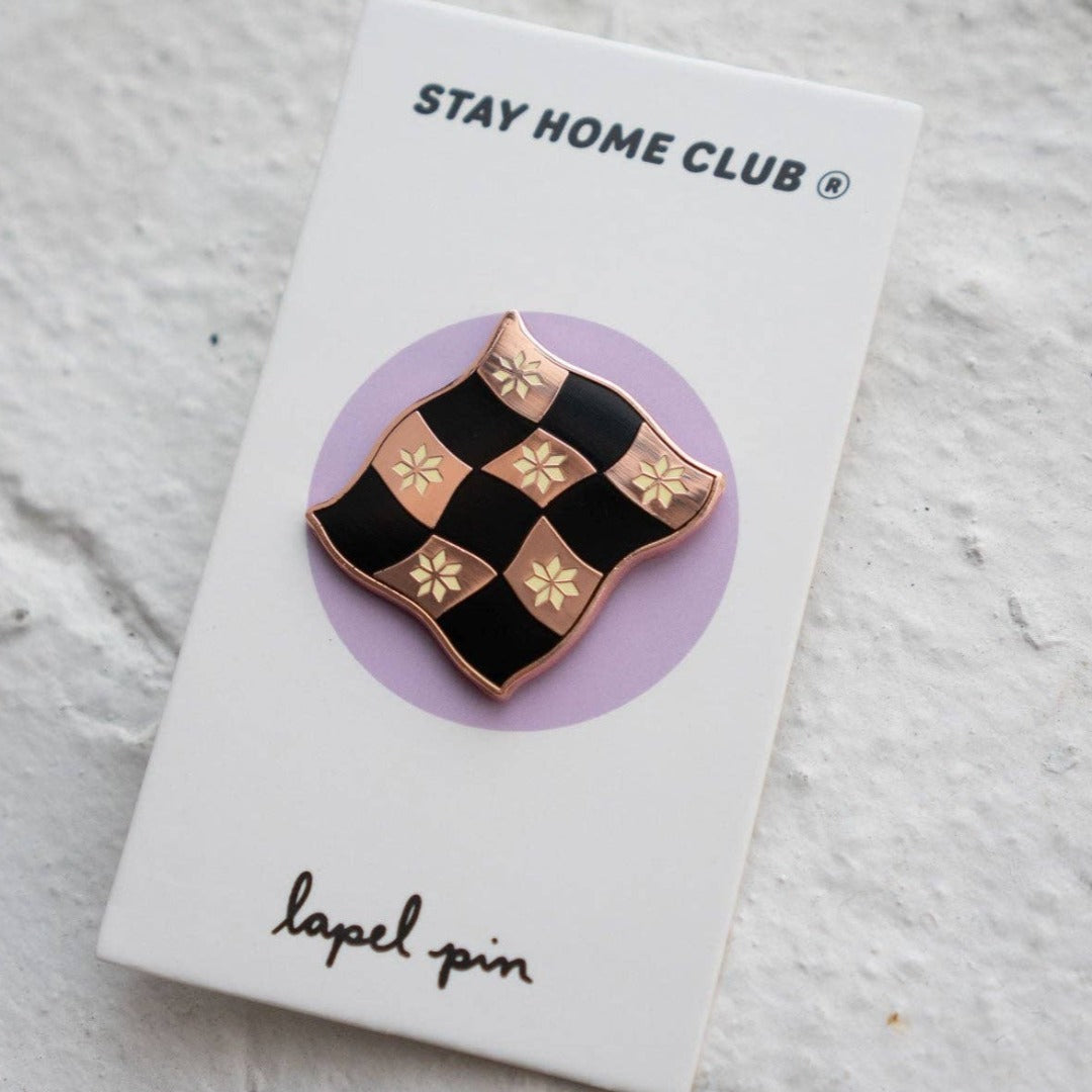 Rose gold-toned pin shaped like a checkered flag with black and white squares. Each square has a small, white flower design in the center.