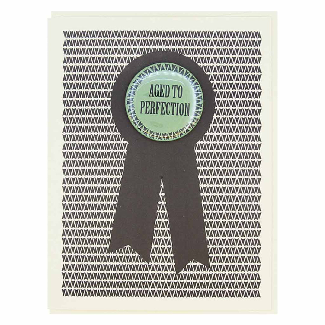 A birthday card with a black and white geometric pattern in the background. A large black ribbon graphic is overlaid, and centered on it is a round, mint green button with a white scalloped edge. The button reads "AGED TO PERFECTION" in black text.