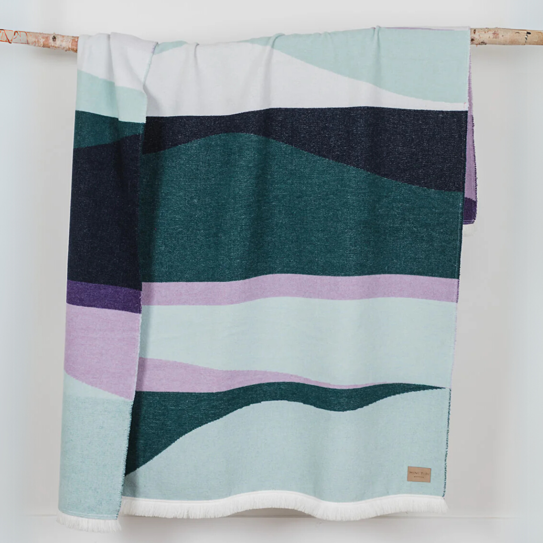 The Aurora Reversible Eco-Friendly Blanket by Mini Tipi, featuring flowing waves of green, purple, black, and white. This reversible and eco-conscious blanket is available in Ottawa, Ontario, Canada.
