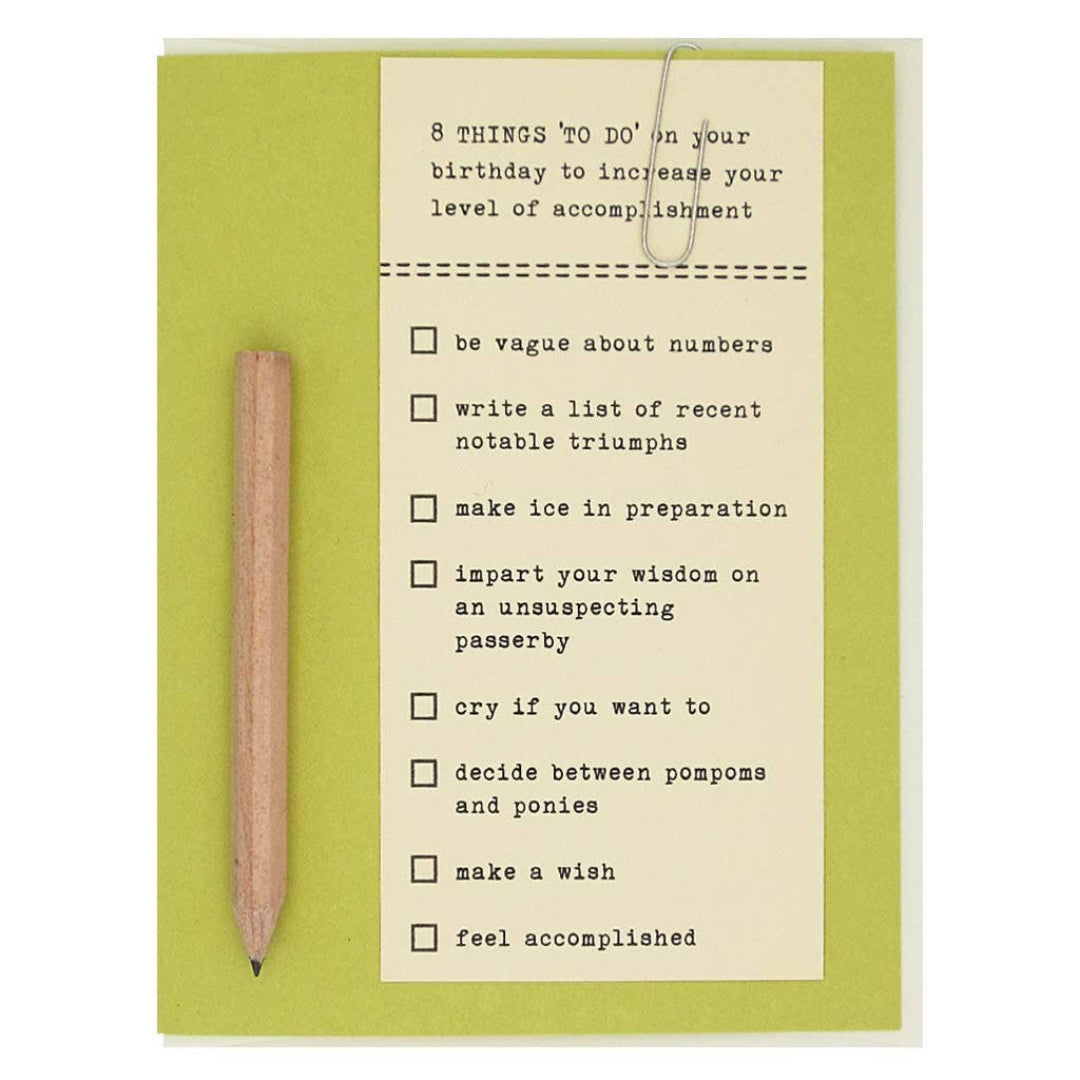 A green and cream-colored card featuring a checklist titled "8 THINGS 'TO DO' on your birthday to increase your level of accomplishment." A small wooden pencil is attached vertically to the left side of the card. The checklist items are "be vague about numbers," "write a list of recent notable triumphs," "make ice in preparation," "impart your wisdom on an unsuspecting passerby," "cry if you want to," "decide between pompoms and ponies," "make a wish," and "feel accomplished."