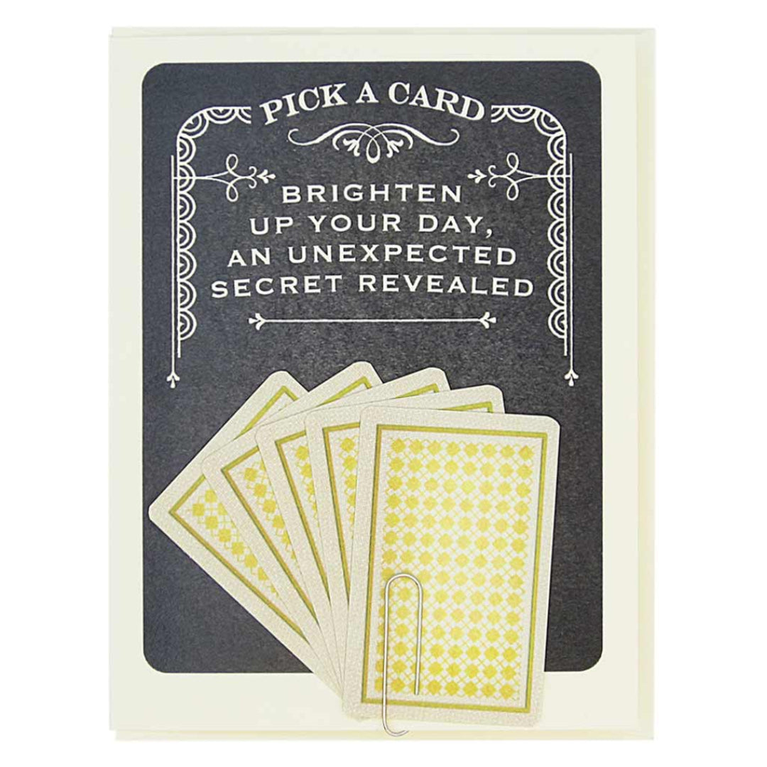 A greeting card with a dark grey background and a decorative white border at the top. The text "PICK A CARD" is at the top, followed by "BRIGHTEN UP YOUR DAY, AN UNEXPECTED SECRET REVEALED." Below the text are five fan-arranged cards with a yellow and white patterned back, held by a paperclip. A second image shows five individual fortune cards, each resembling a playing card with suit symbols and text. The fortunes include messages like "YOUR LUCK IS ABOUT TO CHANGE," "ALL YOUR DREAMS WILL COME TRUE," and "