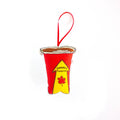A close-up, high-angle flat lay shot on a plain white background features a soft, plush Christmas ornament shaped like a disposable coffee cup. The cup is red with a yellow and red arrow graphic on the front that says "Canada's Favorite" and includes a maple leaf. The top of the cup is brown to resemble coffee and includes tiny text that says "PLEASE FLIP AGAIN / RÉESSAYER". A red ribbon loop is attached to the top for hanging.