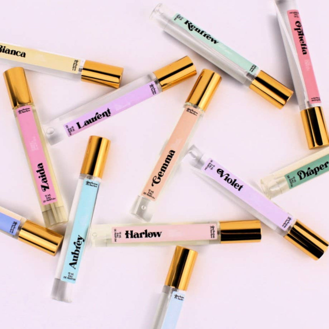A collection of ten small perfume bottles scattered across a white surface. Each bottle has a gold cap and a transparent body, revealing the colored liquid inside. Each bottle has a label with the perfume's name in a playful font: Renfrew, Ophelia, Laurent, Gemma, Violet, Diaper, Bianca, Zaida, Aubrey, and Harlow.