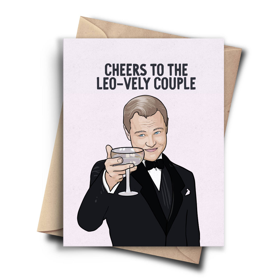 A humorous card featuring a cartoon drawing of Leonardo DiCaprio as Jay Gatsby from The Great Gatsby, holding a cocktail glass and winking. The text on the light pink background reads "CHEERS TO THE LEO-VELY COUPLE." A brown kraft envelope is behind the card.