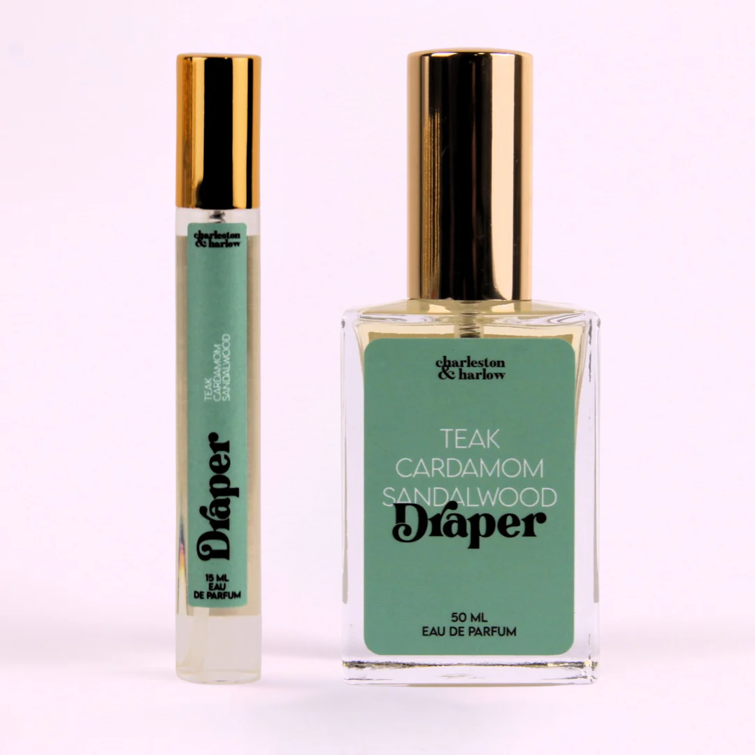A small, cylindrical bottle of perfume with a gold cap. The bottle is transparent and filled with a light-colored liquid. The label on the bottle reads "Draper" in large, black letters. Below the brand name, it lists the fragrance notes: "Teak, Cardamom, Sandalwood."