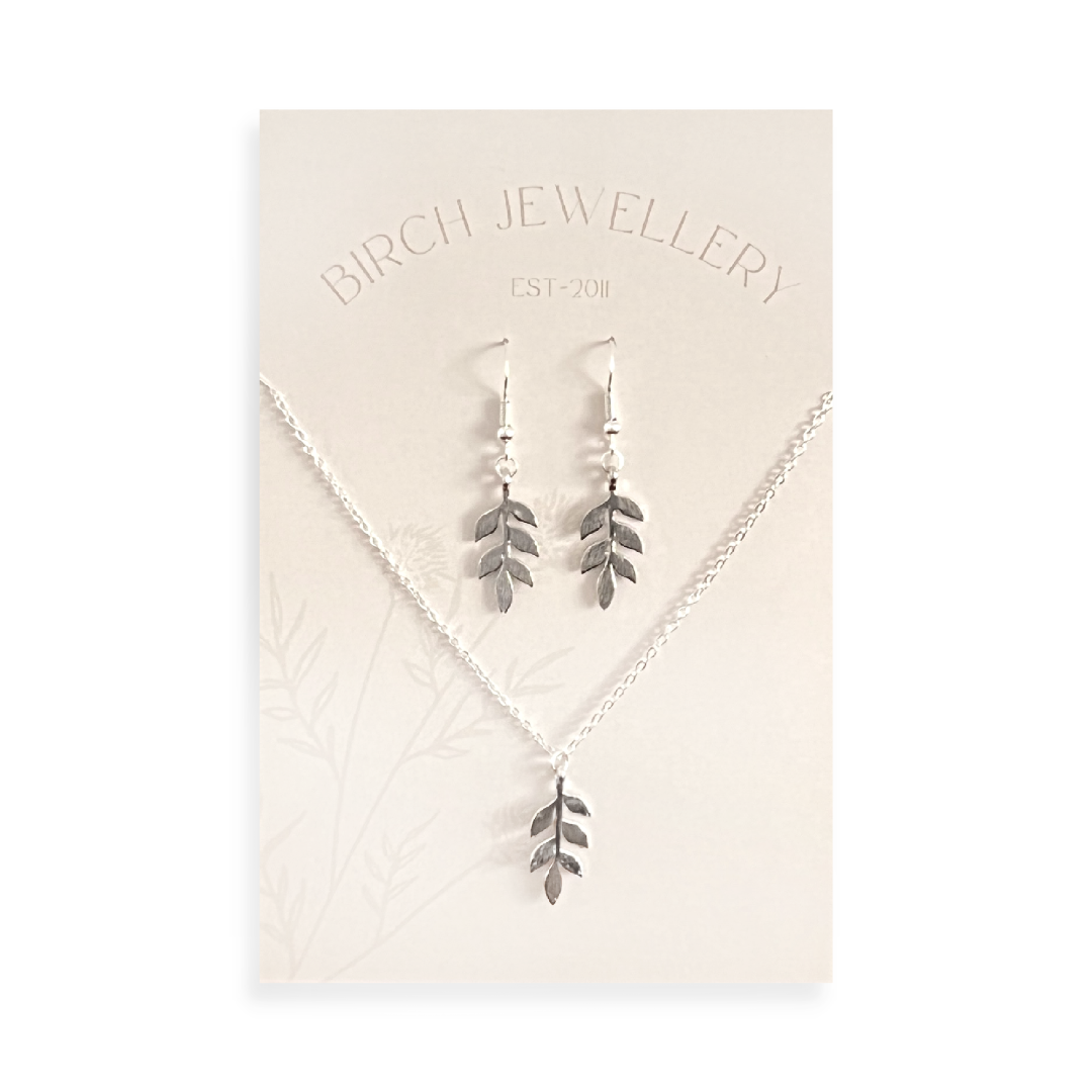 A silver necklace and earring set on a white card. The necklace features a silver leaf pendant, and the earrings are silver leaf-shaped drop earrings.
