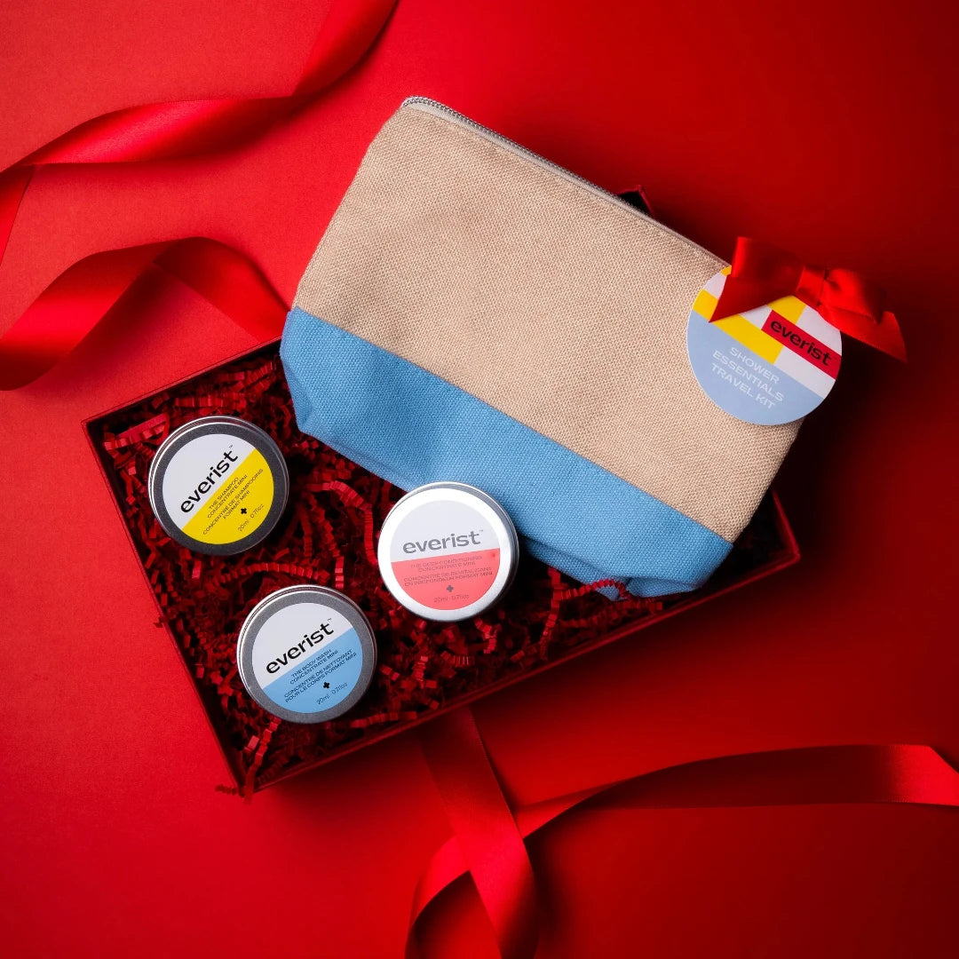 An Everist Shower Essentials Travel Kit. The kit includes a small, blue and beige canvas pouch with a red ribbon and tag that reads "Everist Shower Essentials Travel Kit." Inside the pouch are three small, round tins labeled "Everist" with different colored lids.