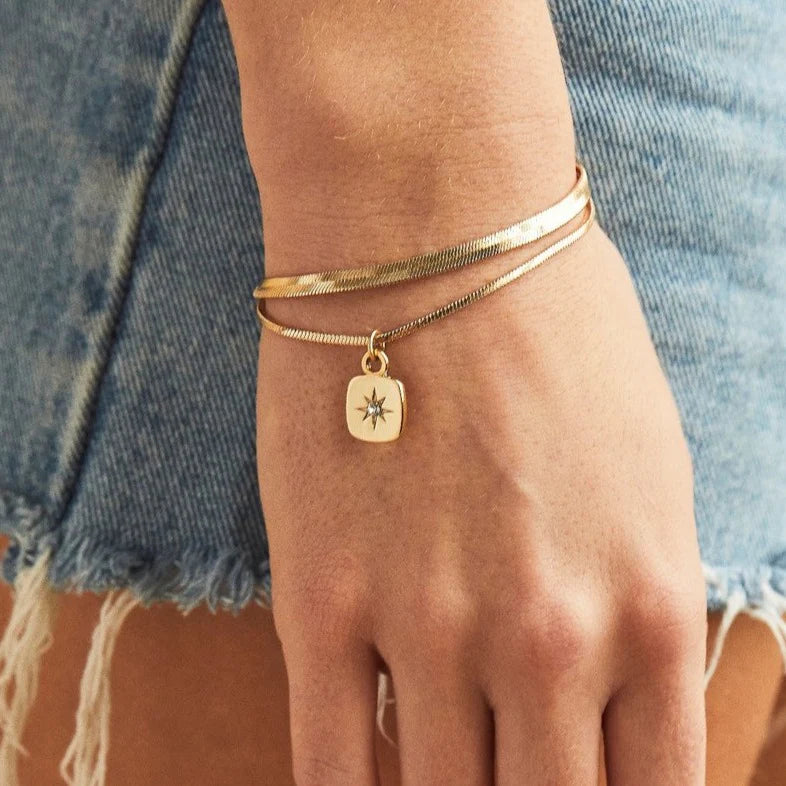 A set of two gold bracelets, one layered over the other, with a star charm hanging from the bottom. The bracelets are displayed on a pink card with the brand name "Foxy Originals."