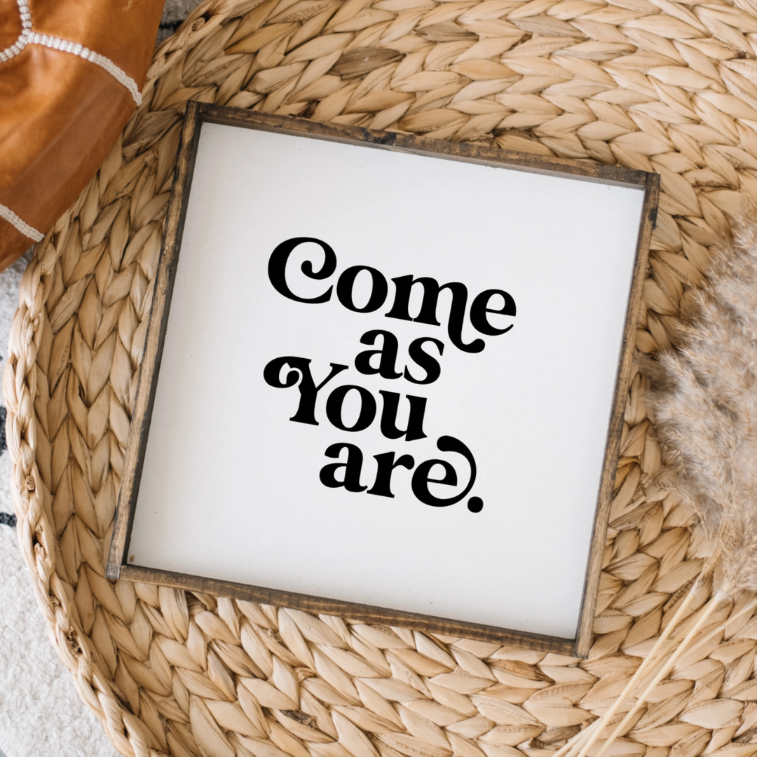 A framed square wood sign with the text "Come as you are" in black, stylized font, centered on a white background. The frame is a dark brown wood with a rustic finish. 