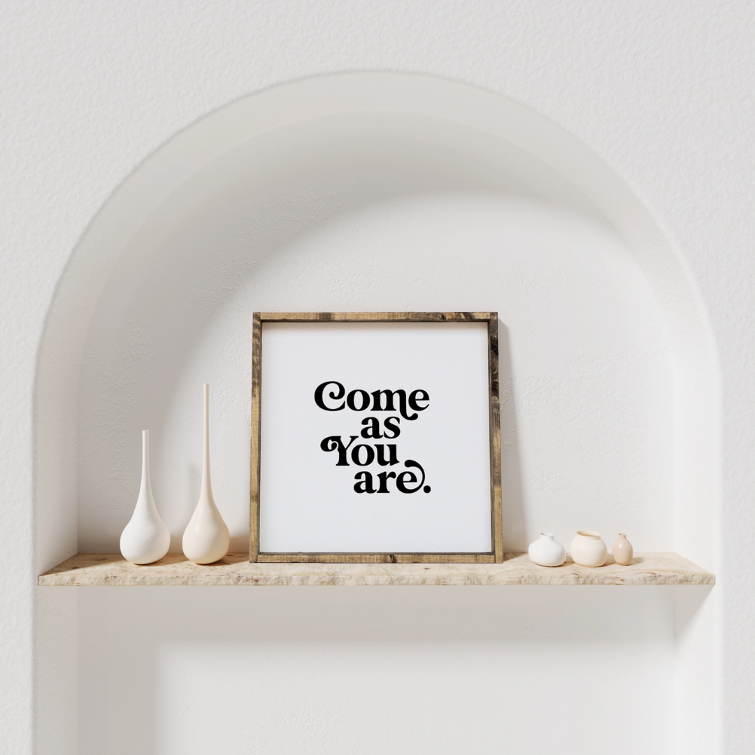 A framed square wood sign with the text "Come as you are" in black, stylized font, centered on a white background. The frame is a dark brown wood with a rustic finish. 