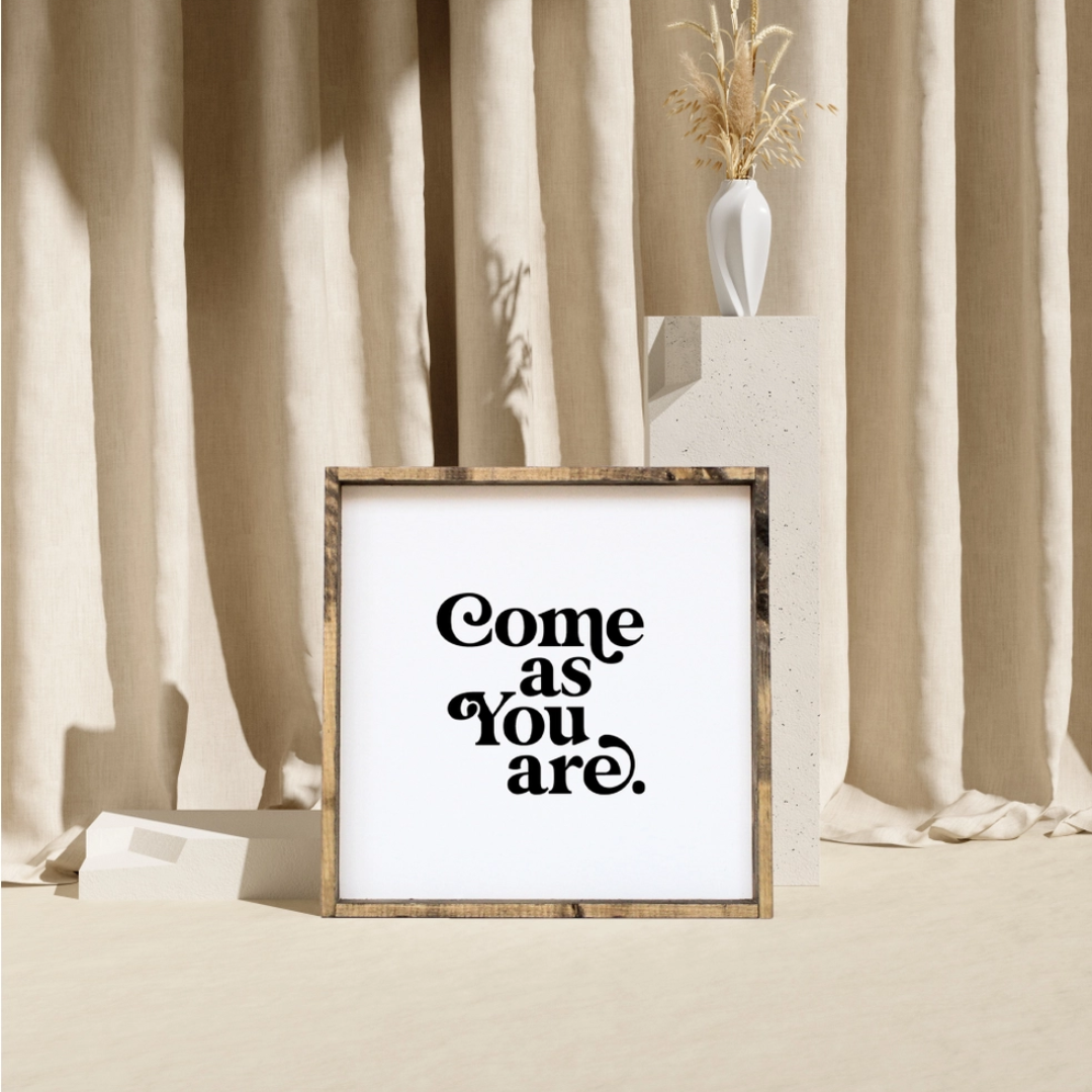 A framed square wood sign with the text "Come as you are" in black, stylized font, centered on a white background. The frame is a dark brown wood with a rustic finish. 
