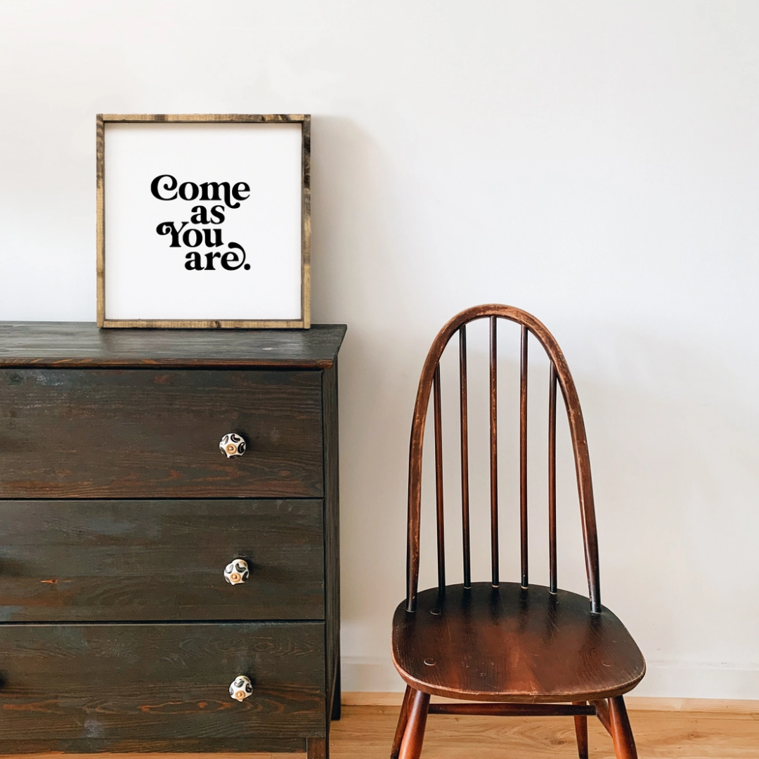 A framed square wood sign with the text "Come as you are" in black, stylized font, centered on a white background. The frame is a dark brown wood with a rustic finish. 