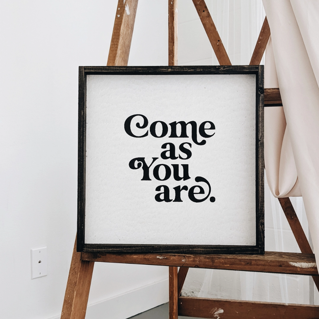 A framed square wood sign with the text "Come as you are" in black, stylized font, centered on a white background. The frame is a dark brown wood with a rustic finish. 