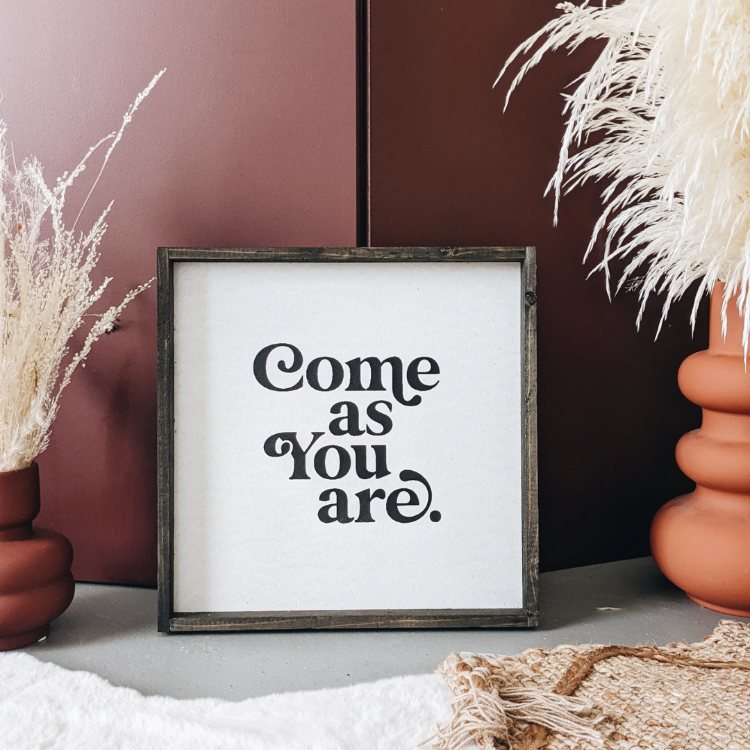 A framed square wood sign with the text "Come as you are" in black, stylized font, centered on a white background. The frame is a dark brown wood with a rustic finish. 