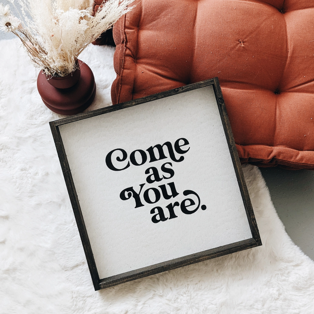 A framed square wood sign with the text "Come as you are" in black, stylized font, centered on a white background. The frame is a dark brown wood with a rustic finish. 
