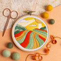 Embroidery hoop with colorful landscape design on a textured surface with yarn and scissors.