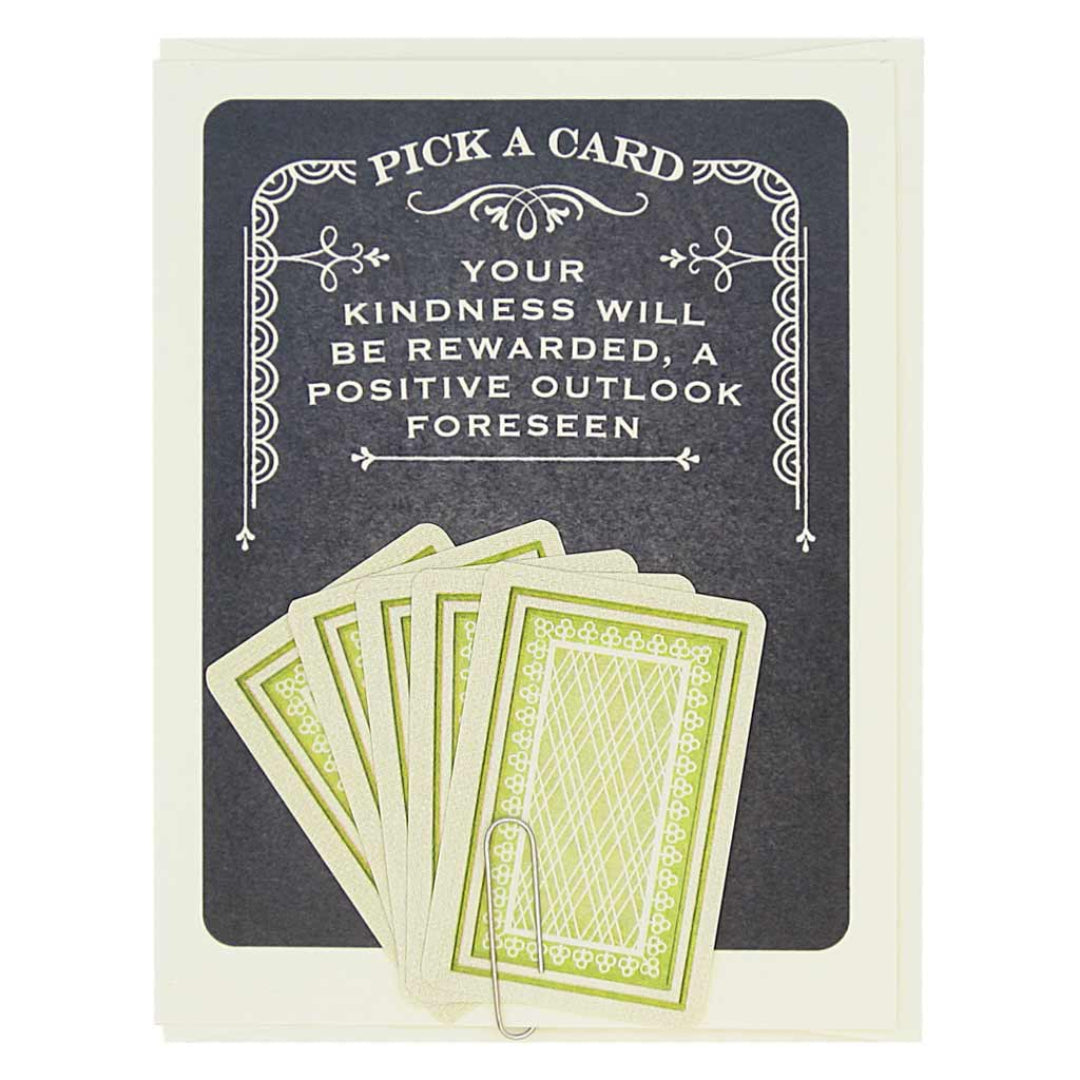 A dark grey card with white decorative borders. The top text reads "PICK A CARD" and below it, "YOUR KINDNESS WILL BE REWARDED, A POSITIVE OUTLOOK FORESEEN". At the bottom, five light green and white playing card-like fortunes are fanned out and held by a paperclip.