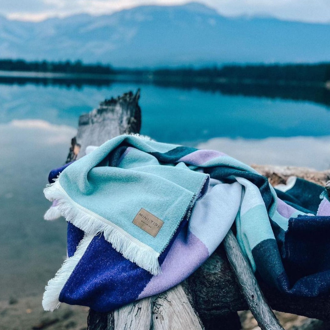 The Aurora Reversible Eco-Friendly Blanket by Mini Tipi, featuring flowing waves of green, purple, black, and white, draped on a natural branch by a lake. This reversible and eco-conscious blanket is available in Ottawa, Ontario, Canada.