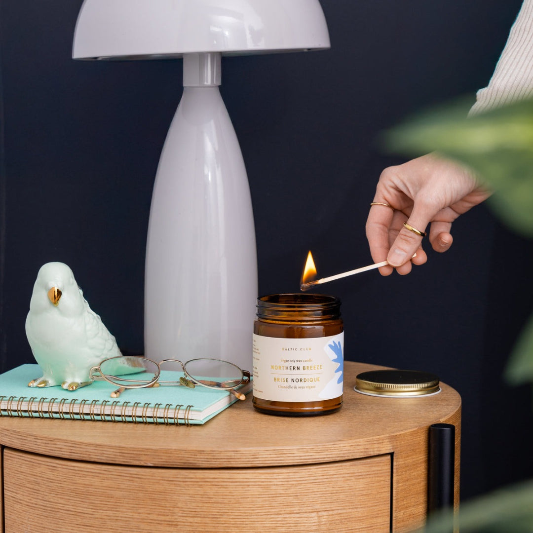 Person lighting a candle on a wooden surface with a lamp and decorative items in the background.