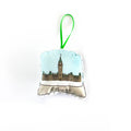 A close-up, high-angle flat lay shot on a plain white background features a soft, plush Christmas ornament shaped like a snow globe. The illustration inside the globe features the Peace Tower of the Canadian Parliament Buildings under a light blue, snowy sky. The base of the globe is white and says "Ottawa" in a black script font. A bright green ribbon loop is attached to the top for hanging.