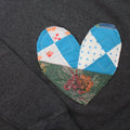 a grey sweatshirt with a large, heart-shaped patch. The patch is made from various fabrics with various patterns, sewn together in a patchwork style.