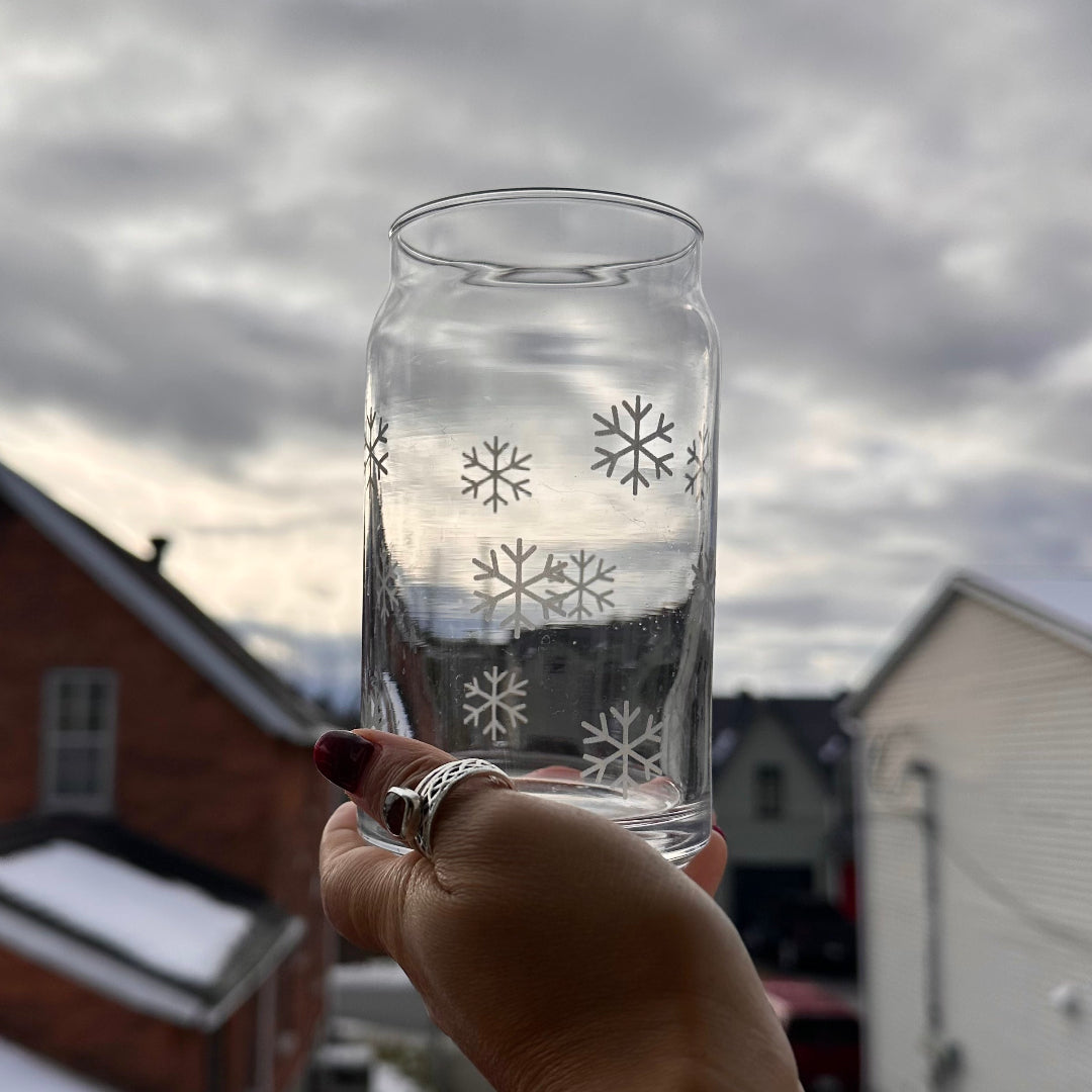 Hand holding a clear glass jar with snowflake designs against a cloudy sky and houses in the background.
