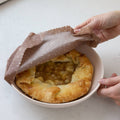 Person covering a baked dish with a brown paper towel on a white surface