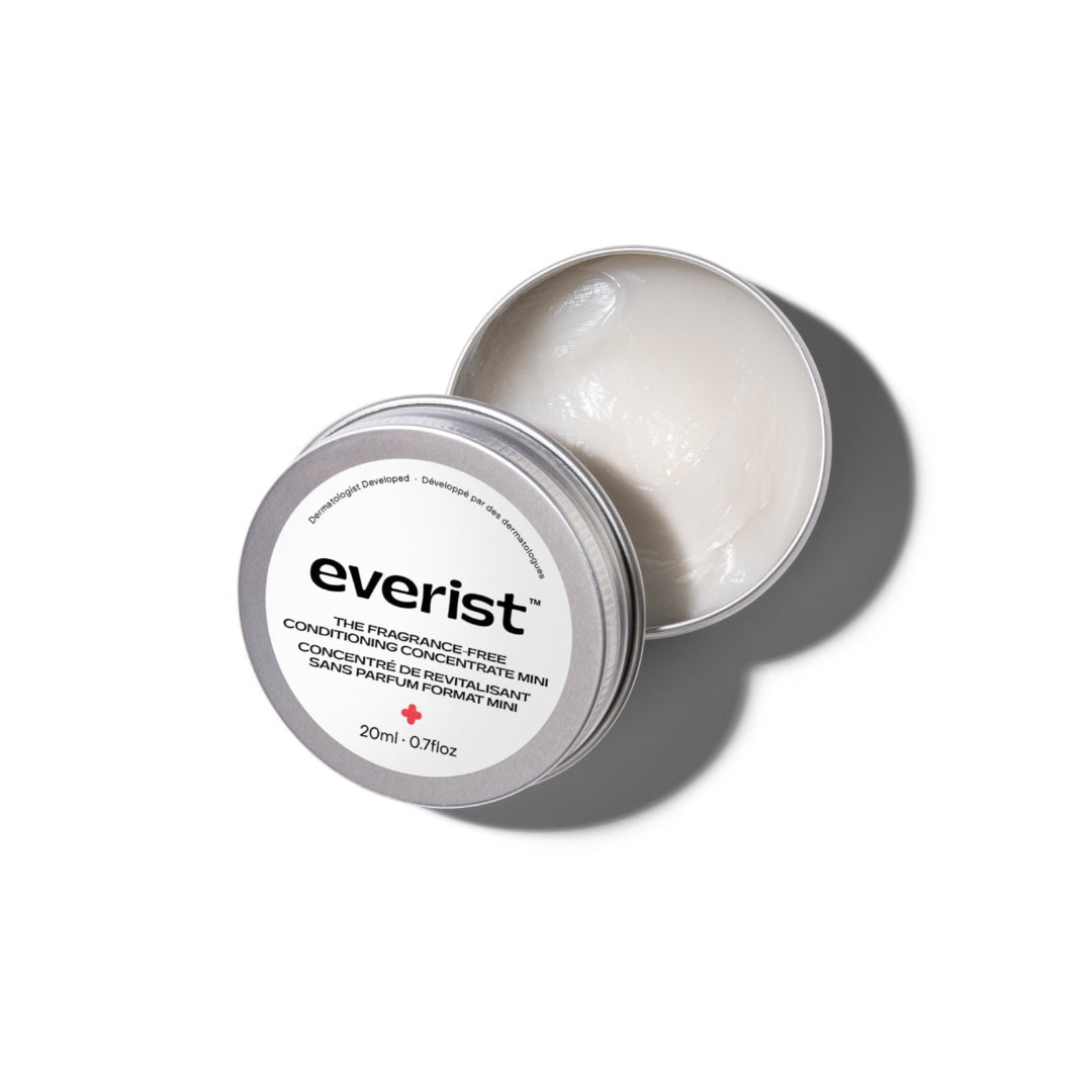 A small, open, silver metal tin of Everist's "The Fragrance-Free Conditioner Concentrate Mini," revealing a translucent, gel-like product inside. The tin's lid is propped against the base, and text on the side indicates it's dermatologist-developed and contains 20ml of product.
