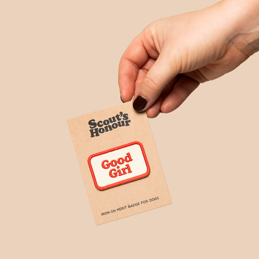 Hand holding a 'Scout's Honour Good Girl' badge on a beige background