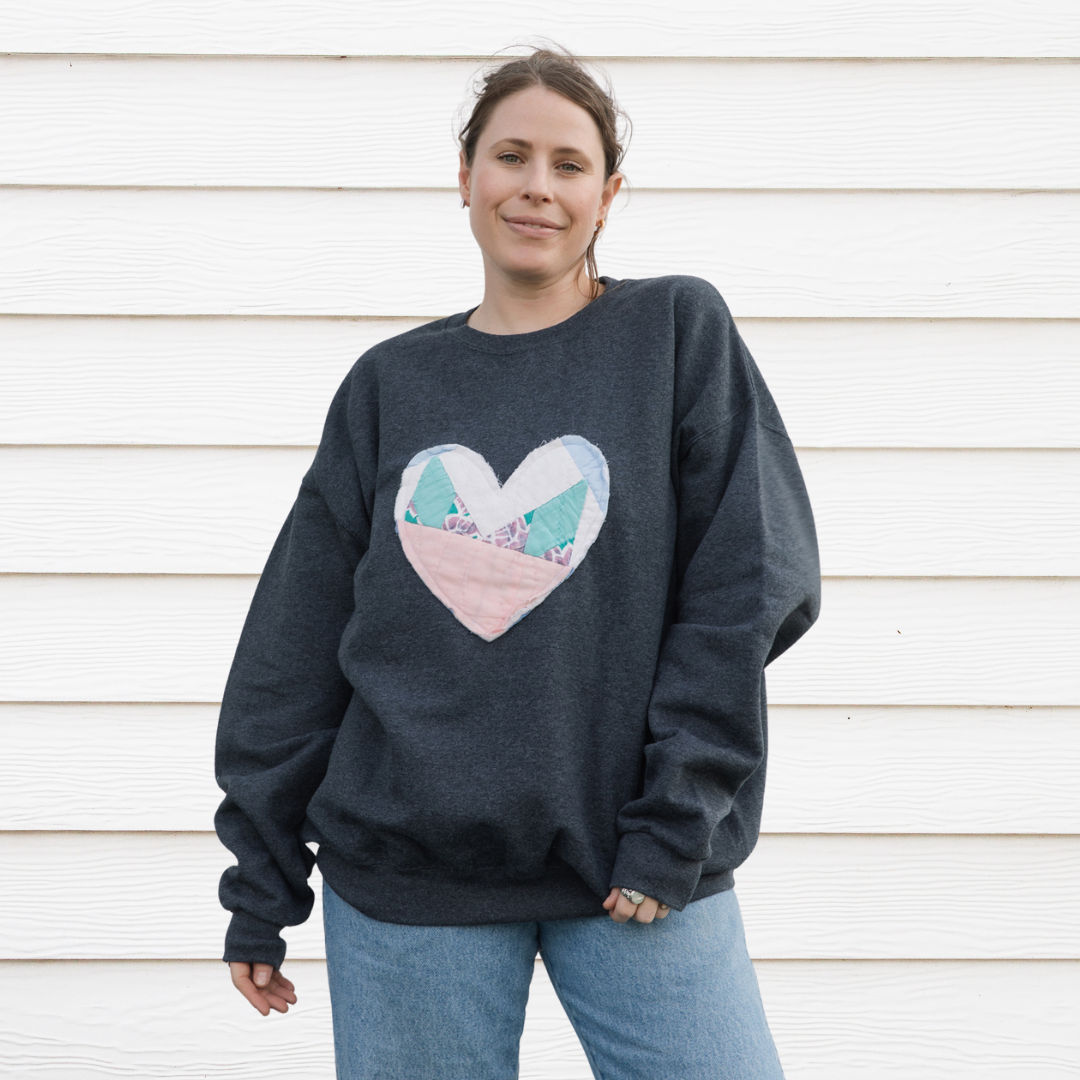 a grey sweatshirt with a large, heart-shaped patch. The patch is made from various fabrics with various patterns, sewn together in a patchwork style.