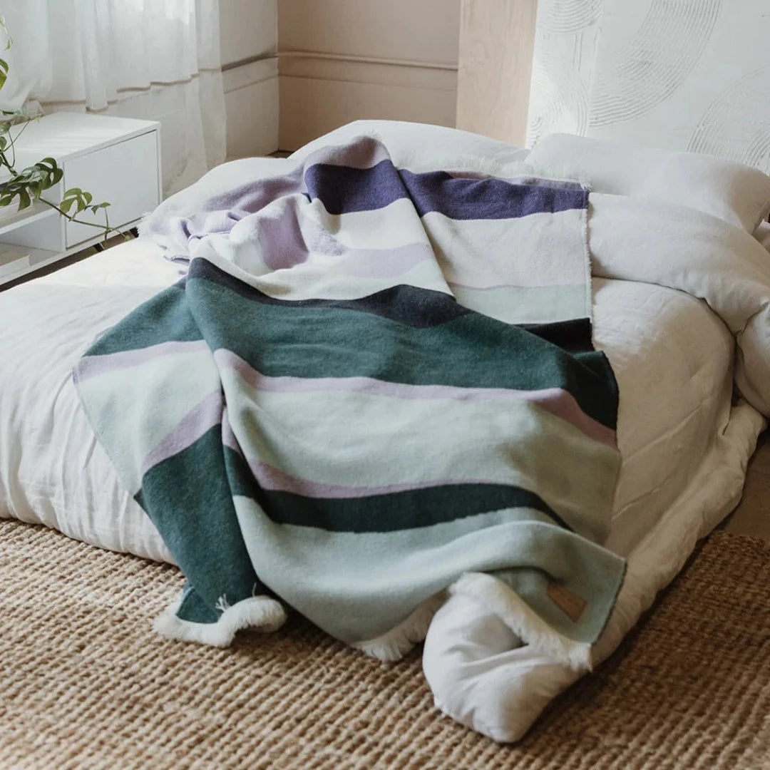 The Aurora Reversible Eco-Friendly Blanket by Mini Tipi, featuring flowing waves of green, purple, black, and white, laying on a bed. This reversible and eco-conscious blanket is available in Ottawa, Ontario, Canada.